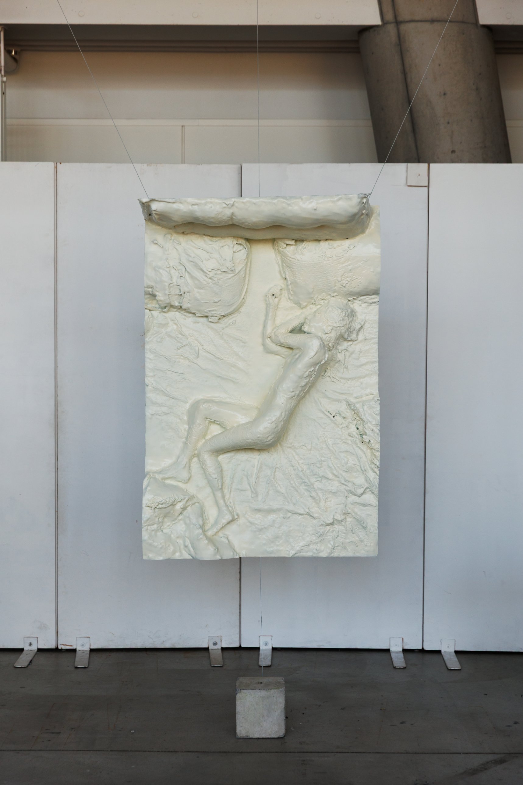 Cream-colored relief of a sleeping figure on a bed, suspended by wires in front of white panels